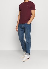 Man in een aansluitend bordeaux shirt, blauwe opgerolde jeans, witte sokken en witte sneakers met rode en zwarte strepen.