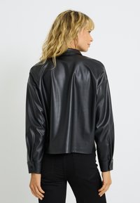 Veste en simili cuir noir avec une coupe décontractée, des épaules tombantes et des détails de poignet boutonnés, présentant une texture lisse et brillante.