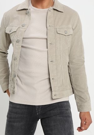 Homme portant une veste en velours côtelé beige sur une chemise côtelée beige clair et un jean gris foncé, debout avec une main dans la poche devant un fond uni.