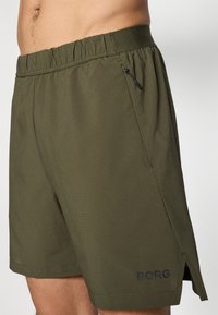 Calções atléticos verdes com um tecido texturizado, cintura elástica e um bolso lateral com fecho e um pequeno detalhe de logótipos.