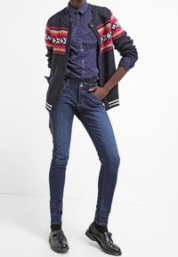 Cárdigan azul marino con un colorido patrón de Fair Isle, camisa oscura, jeans ajustados azul oscuro y zapatos negros. El cárdigan cuenta con botones y ribete a rayas.