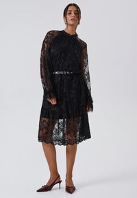 Abito in pizzo nero con maniche lunghe e trasparenti, colletto alto, orlo a balze a strati e dettaglio della cintura metallica. Indossato con tacchi a punta bordeaux.