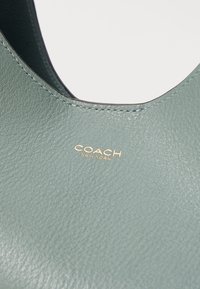 Borsa in pelle verde chiaro in primo piano, caratterizzata da una texture liscia, cuciture visibili e il logo dorato "COACH NEW YORK".