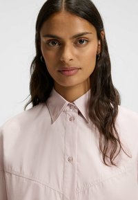 Light pink, oversized shirt with a classic collar, button front, and subtle sheen. Features a chest pocket and textured fabric.