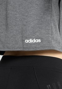 Grå atletisk crop top med texturerad tyg och vit "adidas"-logotyp. Matchas med svarta högmidjade leggings med subtila sömmar.