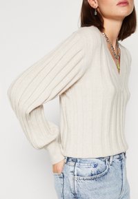 Pull beige côtelé avec un col en V et des manches ballon, fabriqué en matériau doux. Associé à un jean taille haute bleu clair.