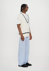Gilet bianco lavorato a maglia con bordo a scallop neri, indossato sopra una t-shirt bianca semplice. I pantaloni a righe blu a gamba larga e mocassini neri completano il look.