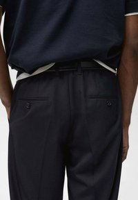 Marineblaues Polohemd mit weißen Akzenten, in maßgeschneiderte schwarze Hose gesteckt. Die Hose hat zwei Gesäßtaschen mit Knopfverschlüssen.