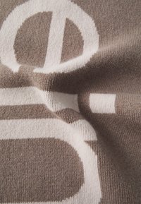 Tessuto a maglia marrone con motivi geometrici bianchi, caratterizzato da un logo circolare prominente. Texture morbida con cuciture e contorni visibili.