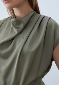 Olive green, draped blouse with soft, smooth fabric; features pleating along the shoulder for texture. Close-up on neckline and sleeve.