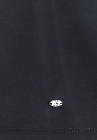 Camiseta de algodón negra con dobladillo cosido y una pequeña placa de logotipo plateada en el centro, añadiendo un detalle sutil a la tela.