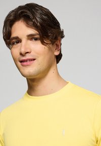 Camiseta amarilla de manga corta, hecha de algodón suave, con cuello redondo y un pequeño logo blanco en la parte izquierda del pecho.