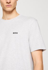 Jeune homme portant un T-shirt gris clair avec le mot "BOSS" imprimé en noir sur la partie gauche de la poitrine.