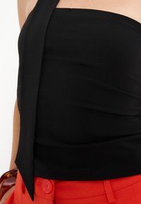 Gros plan d'une personne portant un haut noir à une épaule et un pantalon rouge avec un bouton, montrant une partie de son torse et de son bras.