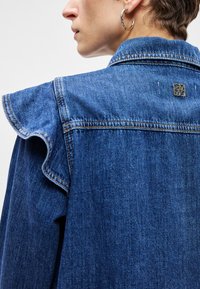 Τζάκετ από τζιν με ρυτιδωμένες ραφές στους ώμους, μπλε ύφασμα, χρυσό έμβλημα στο πίσω μέρος και ορατές λεπτομέρειες ραφής. Υφή, χαλαρός σχεδιασμός.