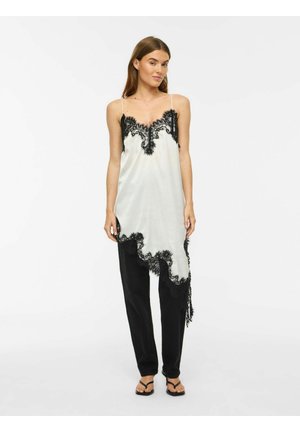 Femeie purtând o rochie albă din satin cu detalii din dantelă neagră peste pantaloni negri și sandale negre, stând pe un fundal alb simplu.