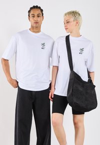 Two models pose together, one wearing a white t-shirt and black jeans, the other in a white t-shirt with a black bag and shorts.