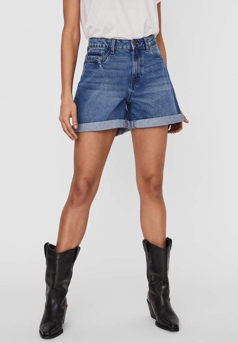 Mujer vistiendo pantalones cortos de denim azul con dobladillo, camiseta blanca metida por dentro, y botas de cowboy negras hasta la mitad de la pantorrilla contra un fondo liso.