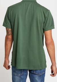 Man som bär en grön kortärmad polo skjorta och blå jeans, stående med ryggen mot betraktaren, visar en tatuering på sin vänstra arm.