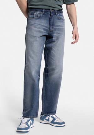 Jeans de mezclilla azul claro con un corte relajado, diseño de cinco bolsillos y un ligero desvanecimiento. Combinados con zapatillas azules y blancas.