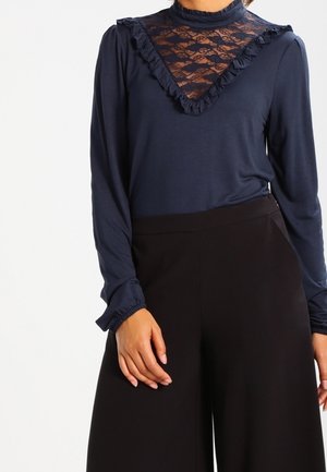 Marineblauw langemouwen top met kantinzet, gerimpelde kraag en manchetten. Gecombineerd met zwarte wijde broek, wat het contrast in texturen en kleuren benadrukt.