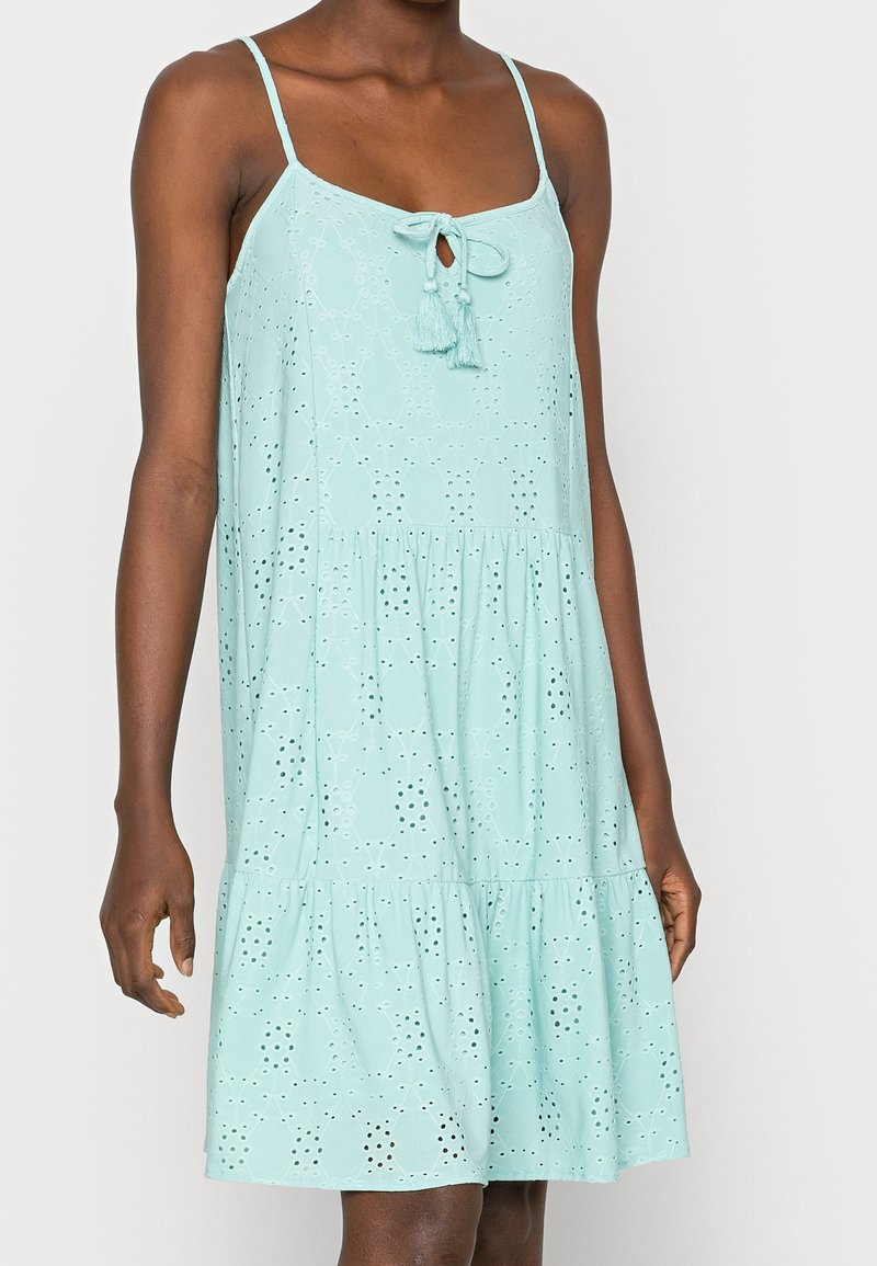 Robe d'été sans manches vert menthe avec motif œillet, jupe à volants, fines bretelles et lien devant avec pompons, portée par une femme.