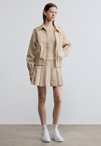 Beige bomberjack met elastische manchetten, gecombineerd met een bijpassende geplooide rok. Witte sneakers en een geribbelde crèmekleurige top maken de look compleet.