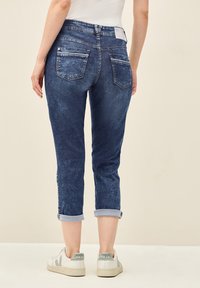 Jean en denim bleu foncé, effilés, avec poches arrière, ourlets roulés et une texture délavée. Associés à des baskets blanches.