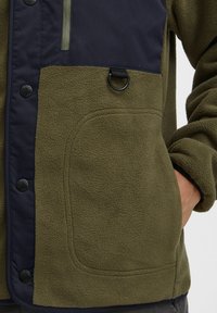 Fleece jacket with olive green body and navy upper section, featuring snap buttons, a zip pocket, and a round black loop detail.