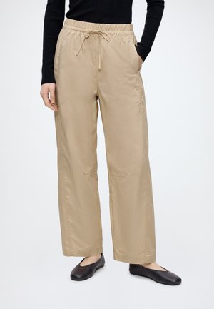 Persoon die beige brede broek met trekkoord in de taille en zwart top met lange mouwen draagt, staand met één hand in de zak en zwarte ballerina's.