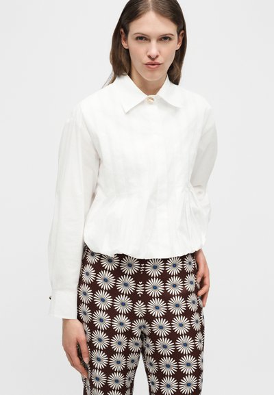 Γυναίκα που φοράει λευκό πουκάμισο με μακριά μανίκια και φαρδιά γιακά, καθώς και καφέ παντελόνι με λευκό λουλουδάτο σχέδιο μαργαρίτας, στέκεται μπροστά σε απλό φόντο.