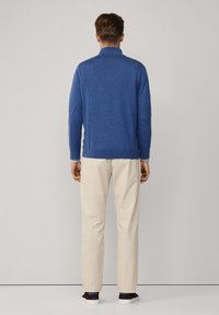 Pull bleu avec col montant, manches longues et poignets côtelés, associé à un pantalon beige et des baskets noires. Texture lisse sur l'ensemble.