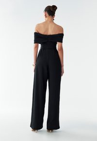 Sort off-shoulder buksedragt med en tætsiddende overdel og vidde benbukser. Tekstureret stof, bælte i taljen, glat detalje på ryggen.