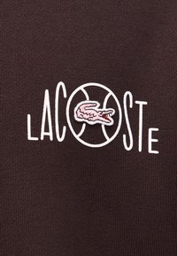 Texte "LACOSTE" avec un contour de ballon de basket blanc remplaçant le "O" et un logo de crocodile brodé rose à l'intérieur du ballon.