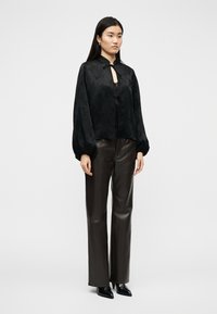 Γυναίκα με μαύρο πουκάμισο με μακριά μανίκια και κορδόνια, σκούρο καφέ δερμάτινο παντελόνι και μαύρες μυτερές γόβες, στέκεται μπροστά σε λευκό φόντο.