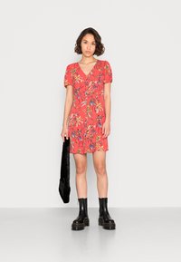 Vestido vermelho de flores, de mangas curtas, com decote em V e detalhes franzidos. Combinado com botas de plataforma pretas até o tornozelo e segurando uma bolsa preta texturizada.