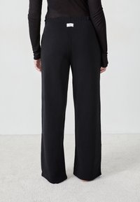 Schwarze weite Stoffhosen mit einem elastischen Bündchen und einem kleinen Etikett auf der Rückseite, die ein sauberes, minimalistisches Design aufweisen.