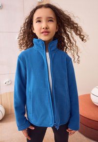 Jachetă fleece albastră cu guler înalt, fermoar integral frontal și buzunare laterale. Textură netedă și croială casual, purtată peste o cămașă albă.