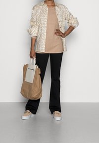 Persona che indossa una camicia a fantasia beige sopra una maglietta color sabbia, pantaloni neri, scarpe slip-on beige, mentre tiene una grande borsa a tracolla beige e bianca.