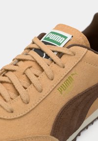 Ίσιο σουέτ αθλητικό παπούτσι σε ταμπά απόχρωση με καφέ λεπτομέρειες, υφή στο επάνω μέρος, επίπεδες κορδόνες, χρυσό λογότυπο και πράσινη ετικέτα. Χαμηλό σχέδιο με κλασική σιλουέτα.