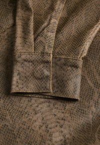 Brūns audums ar čūskas ādas rakstu un teksturētu virsmu. Dizainā redzams režģveida raksts un izteikti salocīts aproces elements.