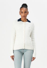 Hvit kabelstrikket cardigan med marineblå krage, knappestolpe og ribbestrikkede mansjetter. Kombinert med lyseblå jeans. Har en detalj med merkevarelogo.