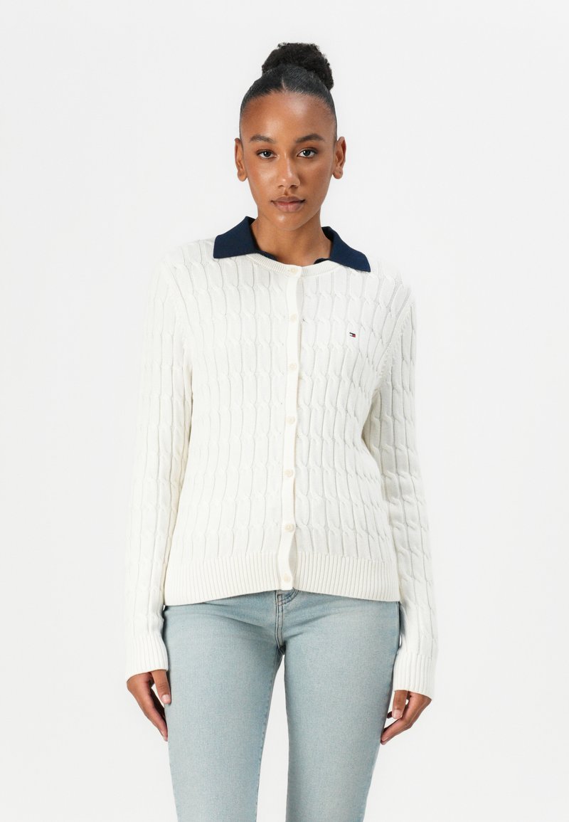 Hvit kabelstrikket cardigan med marineblå krage, knappestolpe og ribbestrikkede mansjetter. Kombinert med lyseblå jeans. Har en detalj med merkevarelogo.