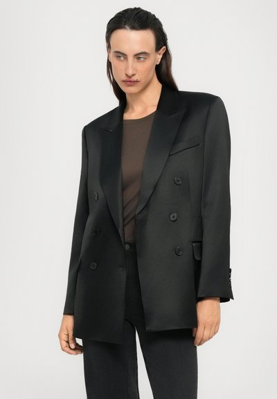 Persoon in een zwart dubbelgebreid blazer over een donkerbruine top met zwarte broek, staand tegen een effen lichte achtergrond.