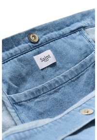 Hele sinine teksakangas väikesega valge sildiga, millel on kiri „Saint Tropez“, mis on õmmeldud metalse kinnitusnupu juurde tasku serval.