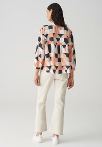 Blusa a maniche lunghe in tessuto a fantasia con forme geometriche nei colori navy, pesca e avorio, abbinata a pantaloni chiari svasati e sneakers bianche.