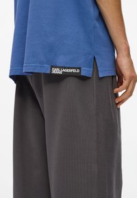 Προφίλ ατόμου που φοράει μπλε πόλο μπλούζα Karl Lagerfeld πάνω από σκούρους γκρι παντελόνες με υφασμάτινη λωρίδα στο πλάι, το χέρι σε χαλαρή θέση στο πλάι.