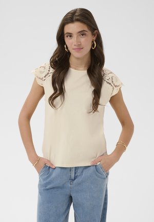 Tânără cu păr lung și castaniu, purtând un top bej cu mâneci din dantelă, blugi albaștri și cercei aurii rotunzi, stând cu mâinile în buzunare.