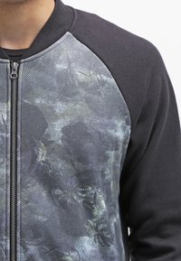 Sudadera negra con cremallera, presenta un frente texturizado y con patrón en tonos de gris y azul, con cuello redondeado y mangas raglán.