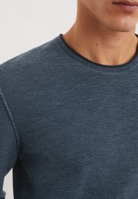Granatowy sweter dzianinowy z okrągłym dekoltem, z teksturowanym wzorem i kontrastującym ciemnym dekoltem. Zbliżenie na górną część ciała.